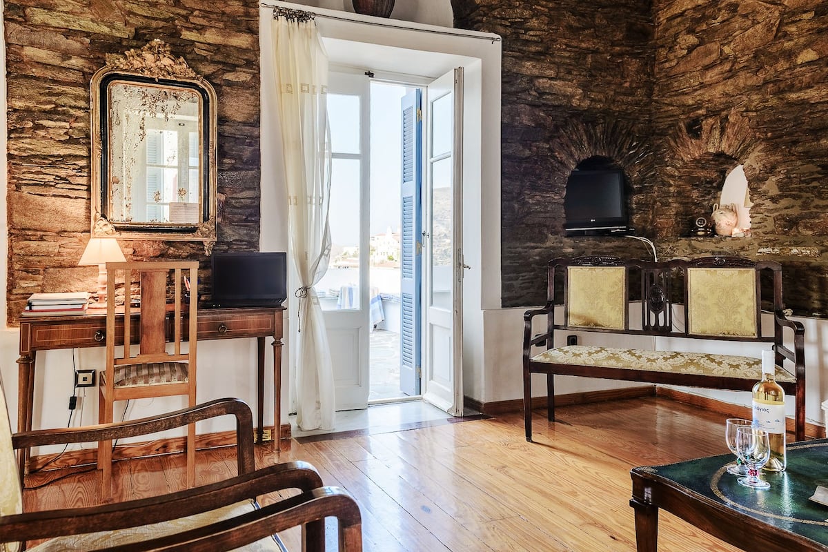 A room with a stone wall, a wooden desk with a television, a chair, and a table with a bottle and glasses.