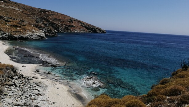 A coastal landscape with a clear blue sea, rocky shore, and a hilly backdrop.