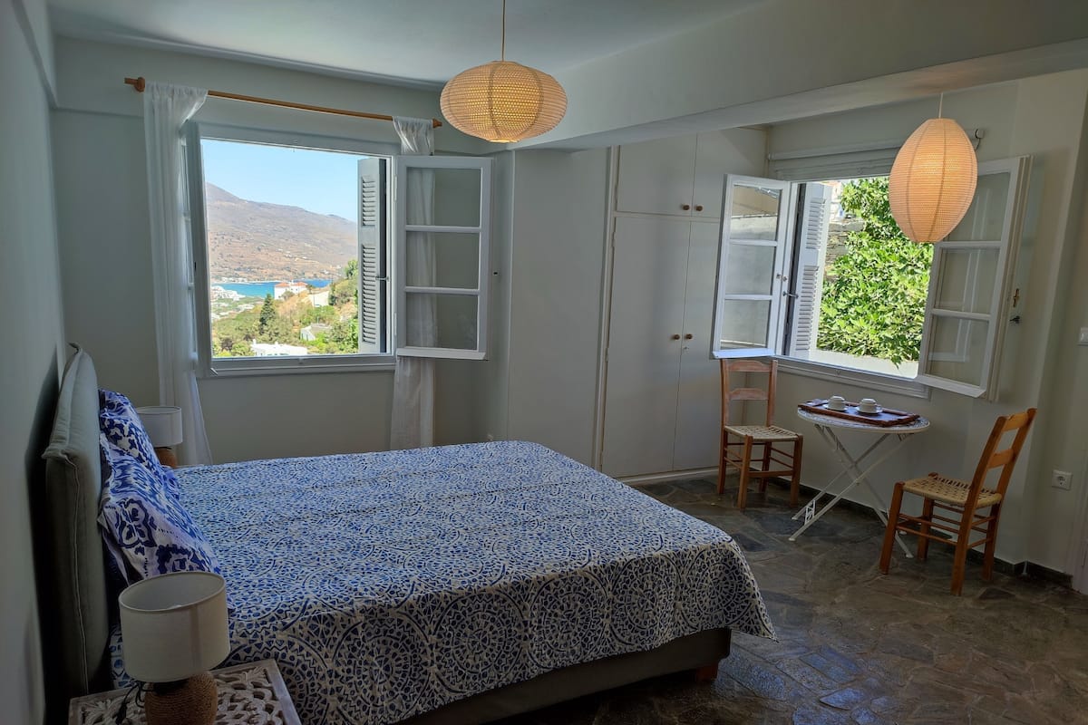 A bedroom with a bed, two windows, a chair, a small table, and a lamp.