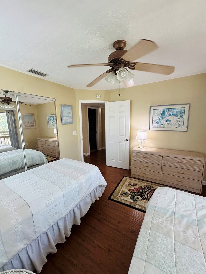 A bedroom with two beds, a ceiling fan, a dresser, and a door leading to another room.