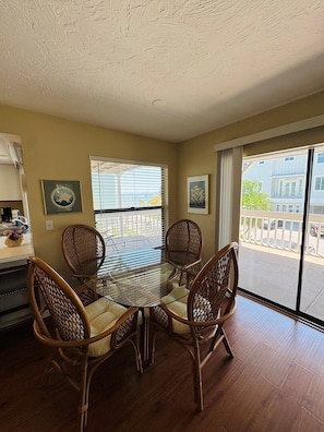A dining area with a glass table and wicker chairs, a kitchen area with a counter and appliances, and a view of a balcony with a railing.