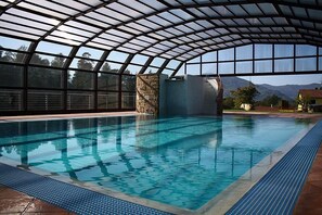 Una piscina interior con techo de vidrio, pilares de piedra y vistas a las montañas.