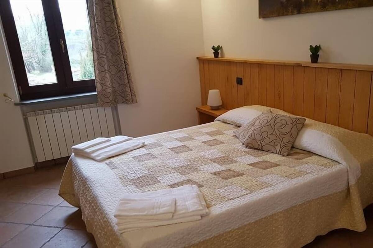 Una cama bien tendida con colcha, dos lámparas de noche y una ventana con cortinas.