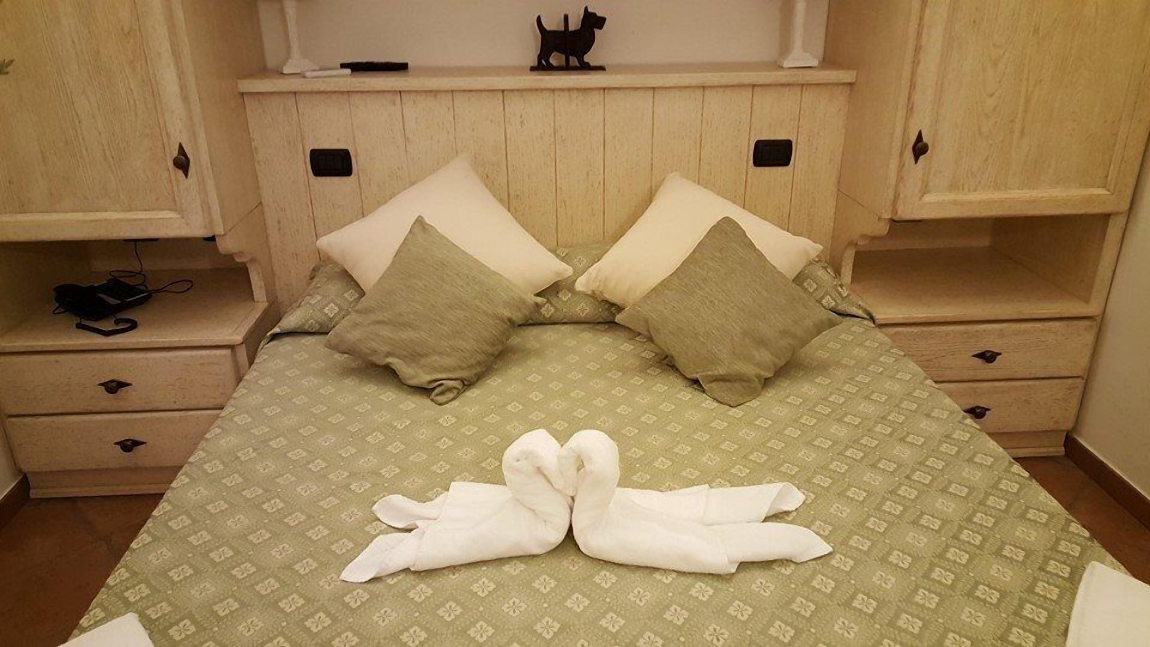 A neatly made bed with a green patterned bedspread and two pillows, a wooden nightstand with a black object on it, and a small shelf above the bed with a dog-shaped decoration.