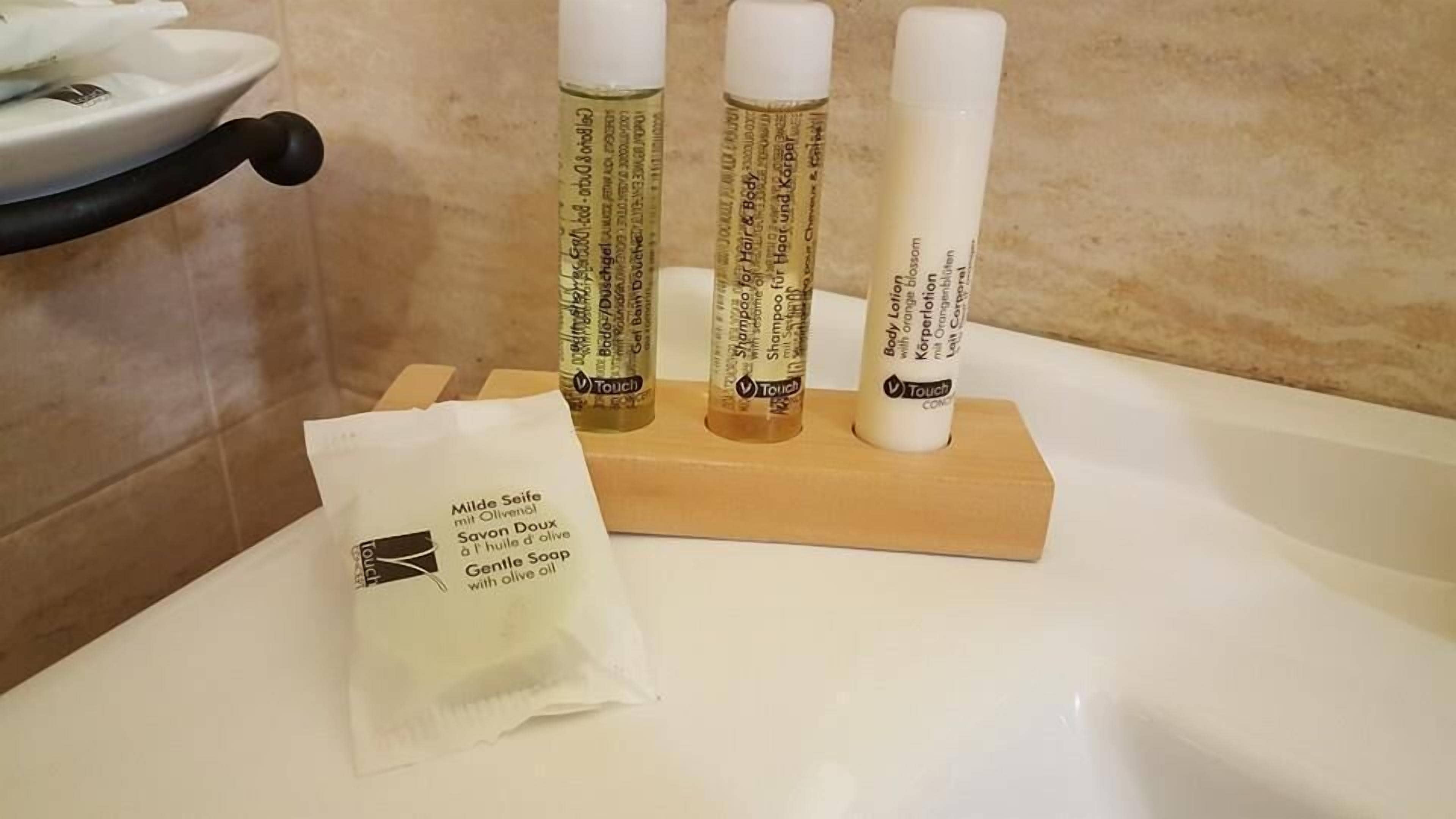 Casa de banho com uma banheira branca, um tabuleiro de madeira com três frascos de loção corporal Touch e um saquinho de sabonete Gentle Soap.