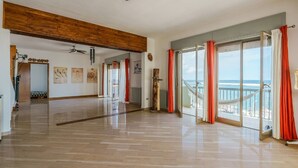 A spacious room with a polished wooden floor, a ceiling fan, and a large sliding glass door leading to a balcony with a view of the sea.