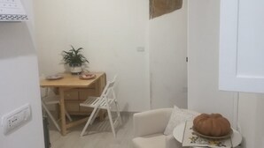 A small, well-lit room with a wooden dining table, a white chair, a small white sofa, and a white side table with a decorative item.