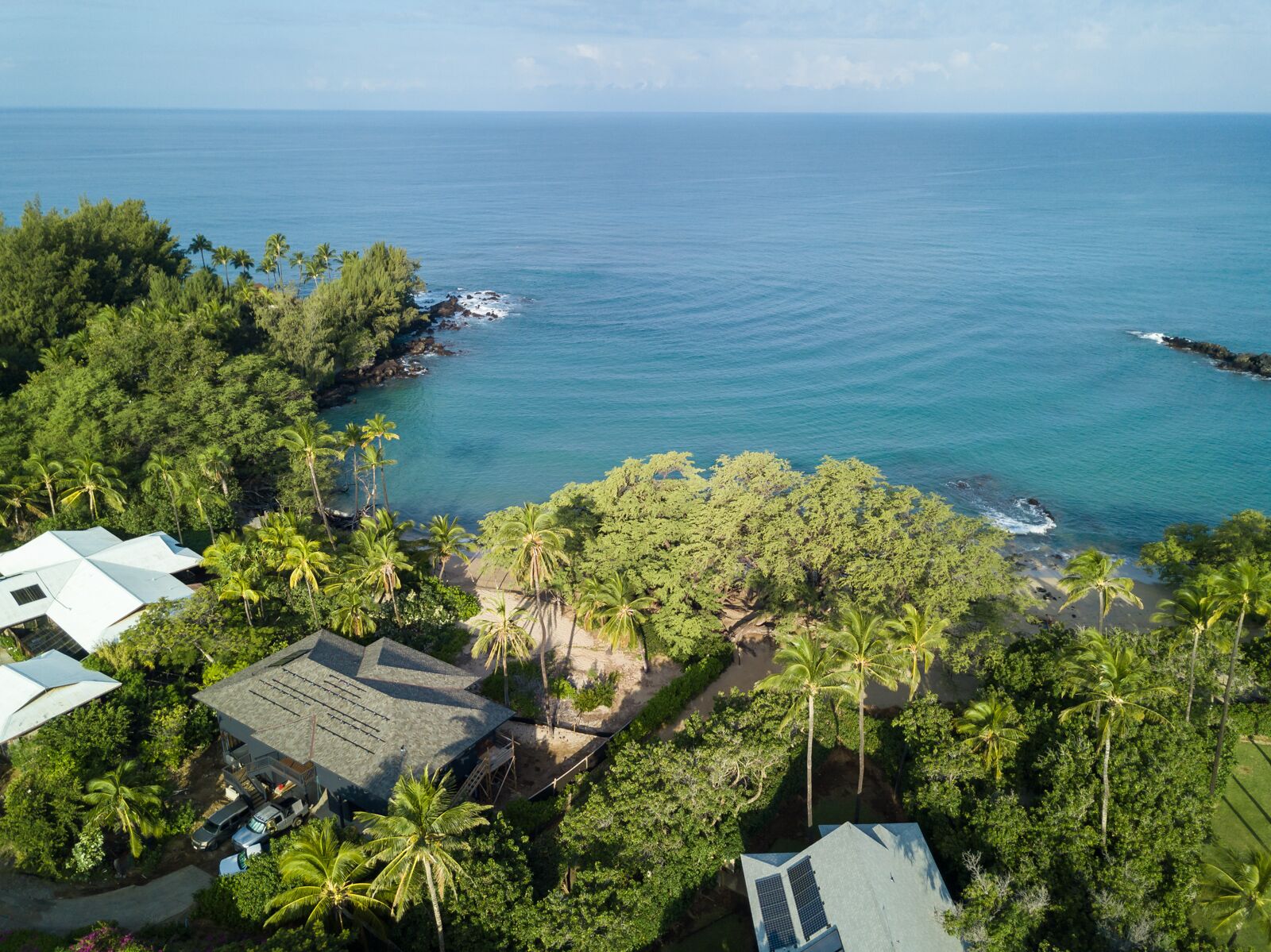 An aerial view of a coastal property with a house, a small dock, and a beach area surrounded by dense tropical vegetation.