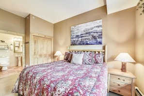 A bedroom with a large bed, wooden headboard, two bedside tables with lamps, a wooden wardrobe, a bathroom with a toilet and sink, and a painting on the wall.