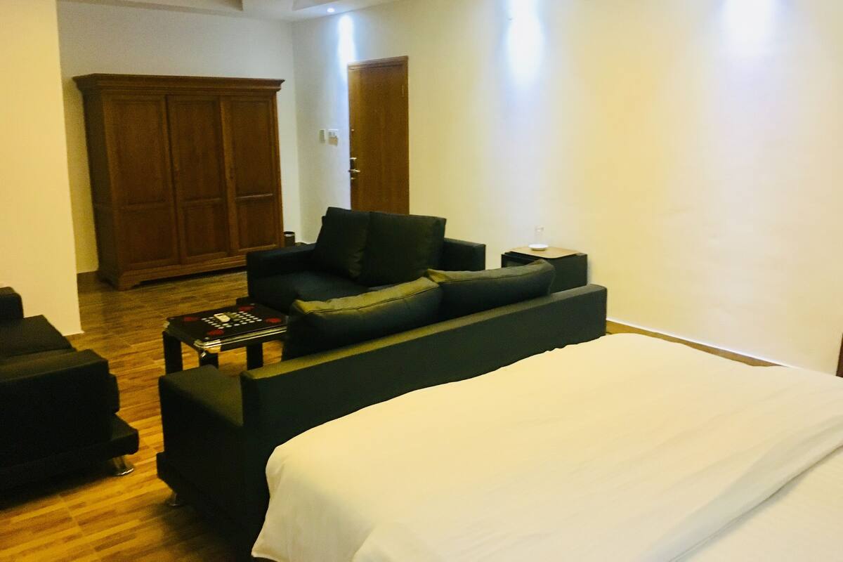 Una habitación de hotel moderna con una cama grande, un sofá oscuro, una pequeña mesa de centro y un armario de madera.