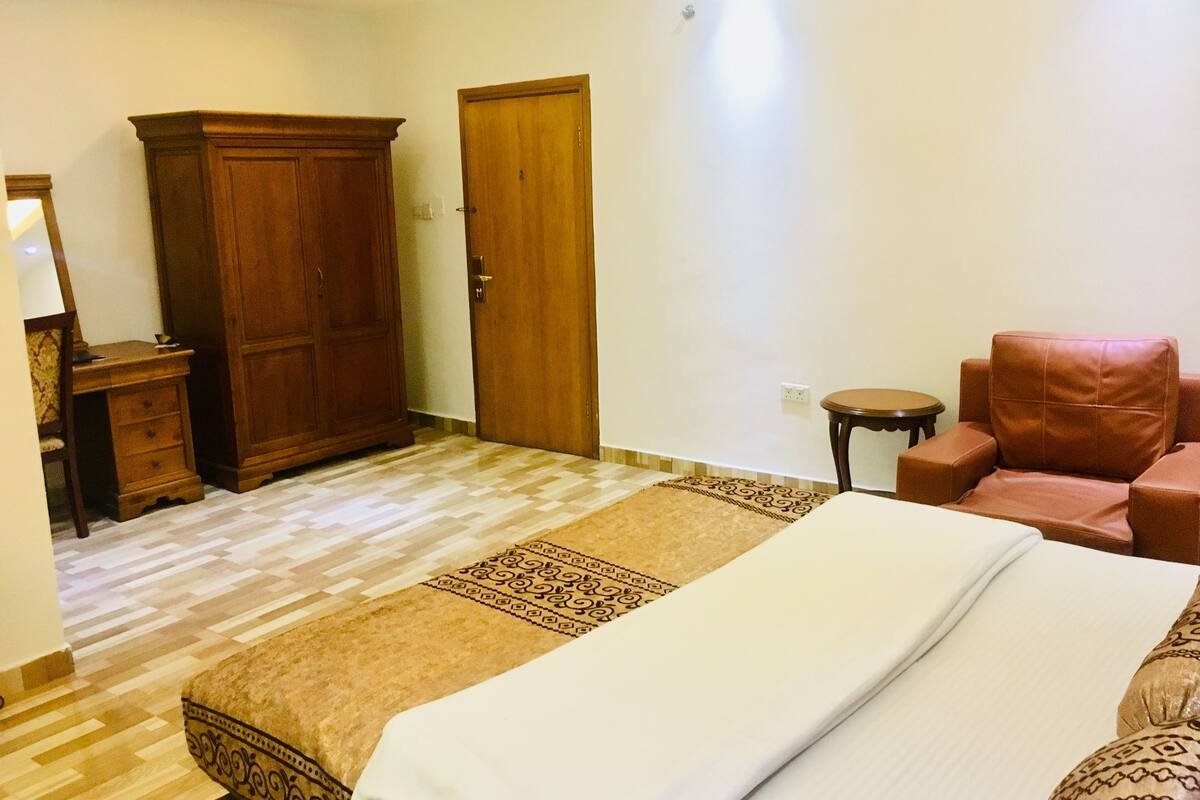 Habitación de hotel con una cama, un armario de madera, un sillón de cuero marrón, una mesita redonda y un tapete estampado.