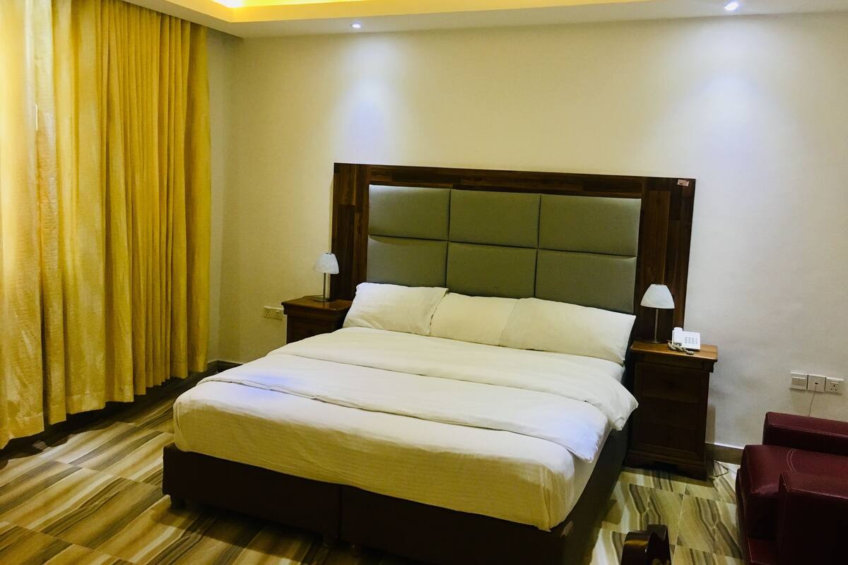 Una habitación de hotel con una cama grande, cabecera de madera, dos mesitas de noche con lámparas, una silla roja y una cortina.