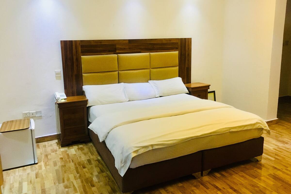 Una cama bien tendida con un cabecero de madera, una mesita de noche con lámpara y un pequeño armario blanco.