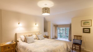 A bedroom with a large bed, bedside tables, a window with curtains, a chair, and wall-mounted lights. - Cuckoo Tor - Two Bedroom Cottage, Sleeps 4 (Lanteglos)