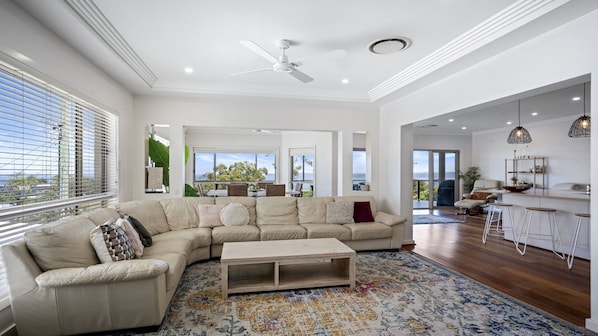 A spacious living room with a large L-shaped sofa, a coffee table, and a patterned rug. There is a ceiling fan, a kitchen area with bar stools, and large windows offering a view of the outdoors.
