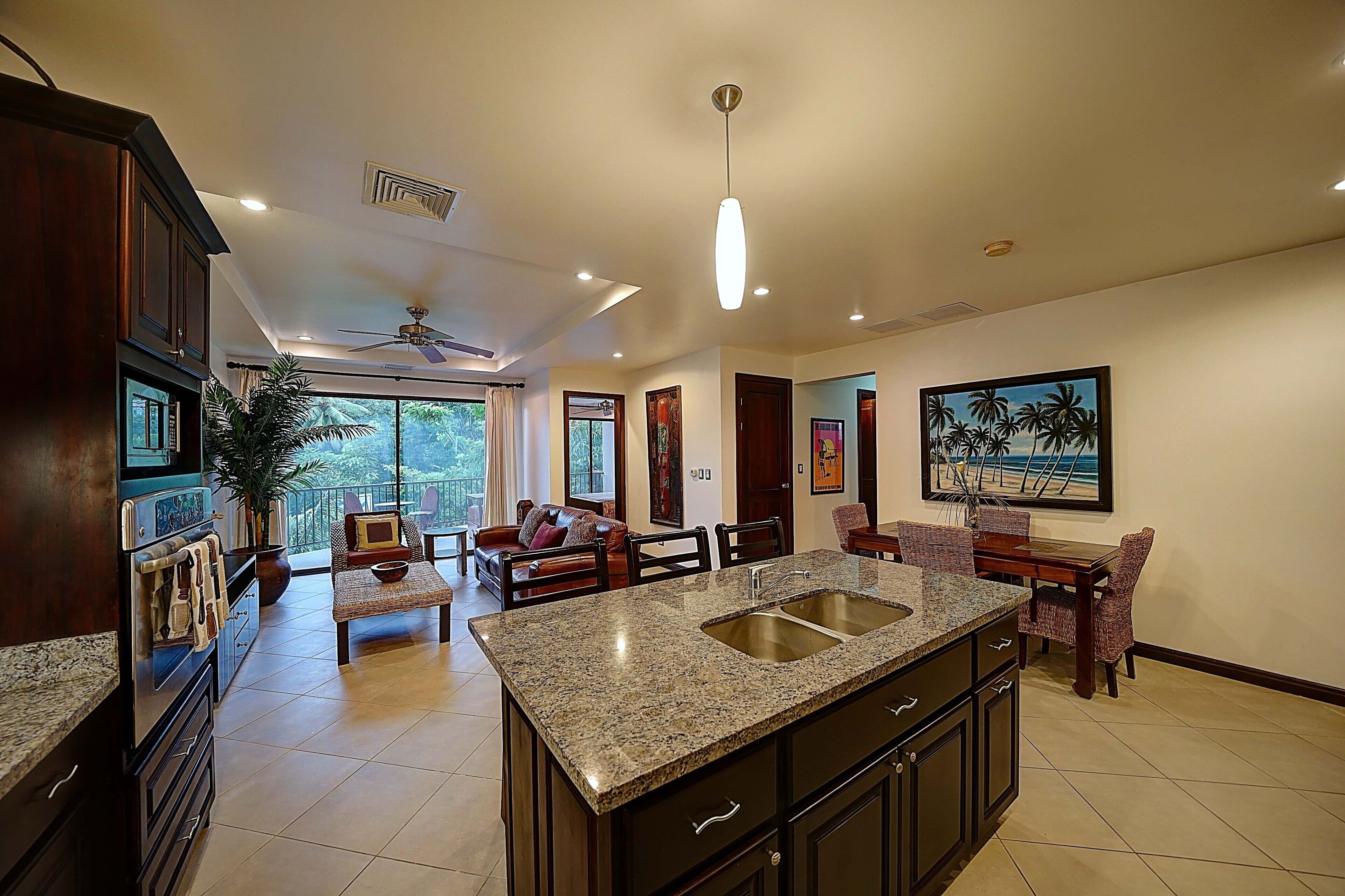 Cocina amplia con encimeras de granito, una isla central con lavamanos y armarios oscuros. Cuenta con un comedor con mesa y sillas, un ventilador de techo y un ventanal con vistas al exterior.