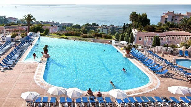 A large outdoor swimming pool with multiple lounge areas and umbrellas, surrounded by buildings and a view of the sea. - 2-room apartment with terrace 800 meters from the beaches (CANNES)