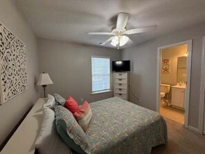 A bedroom with a bed, a ceiling fan, a TV, and a bathroom visible through an open door. - Summer's Den 5 Bedroom Home by RedAwning (Surfside Beach)