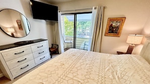 Een slaapkamer met een bed, een dressoir, een televisie, een stoel, een raam met gordijnen en een schilderij aan de muur.