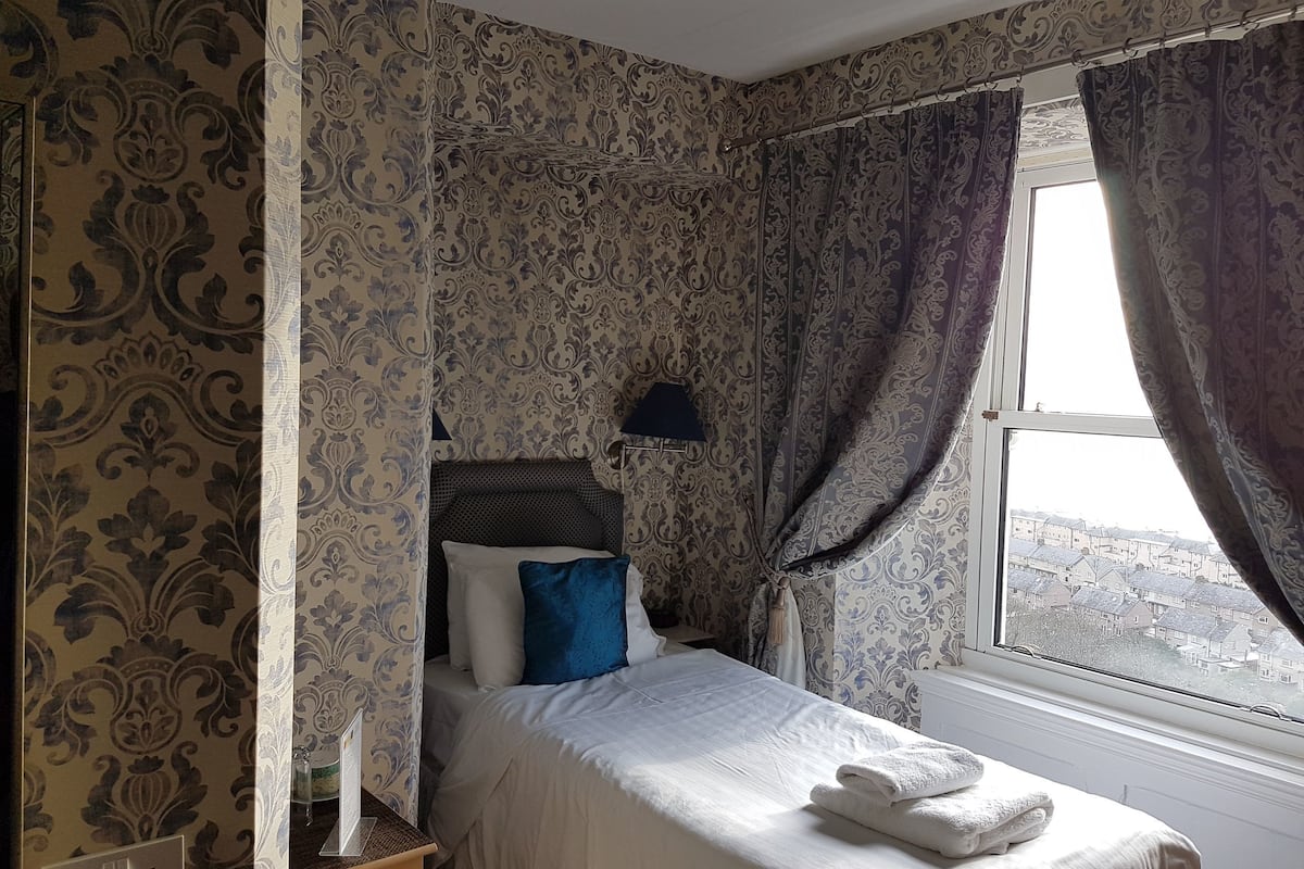 A bedroom with patterned wallpaper, a bed with white bedding and blue pillows, a bedside lamp, and a window with curtains.
