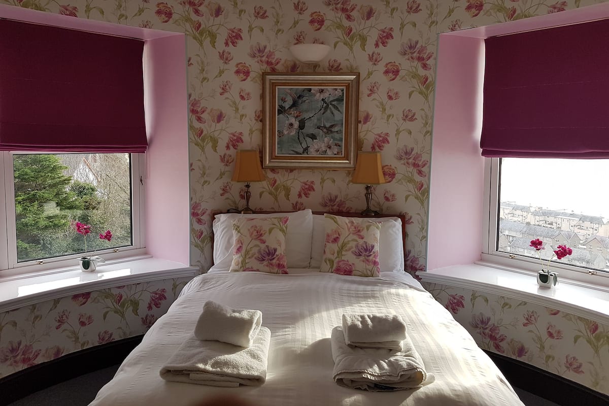 A hotel room with floral wallpaper, a bed with white linens and two towels, two bedside lamps, and a painting above the bed.