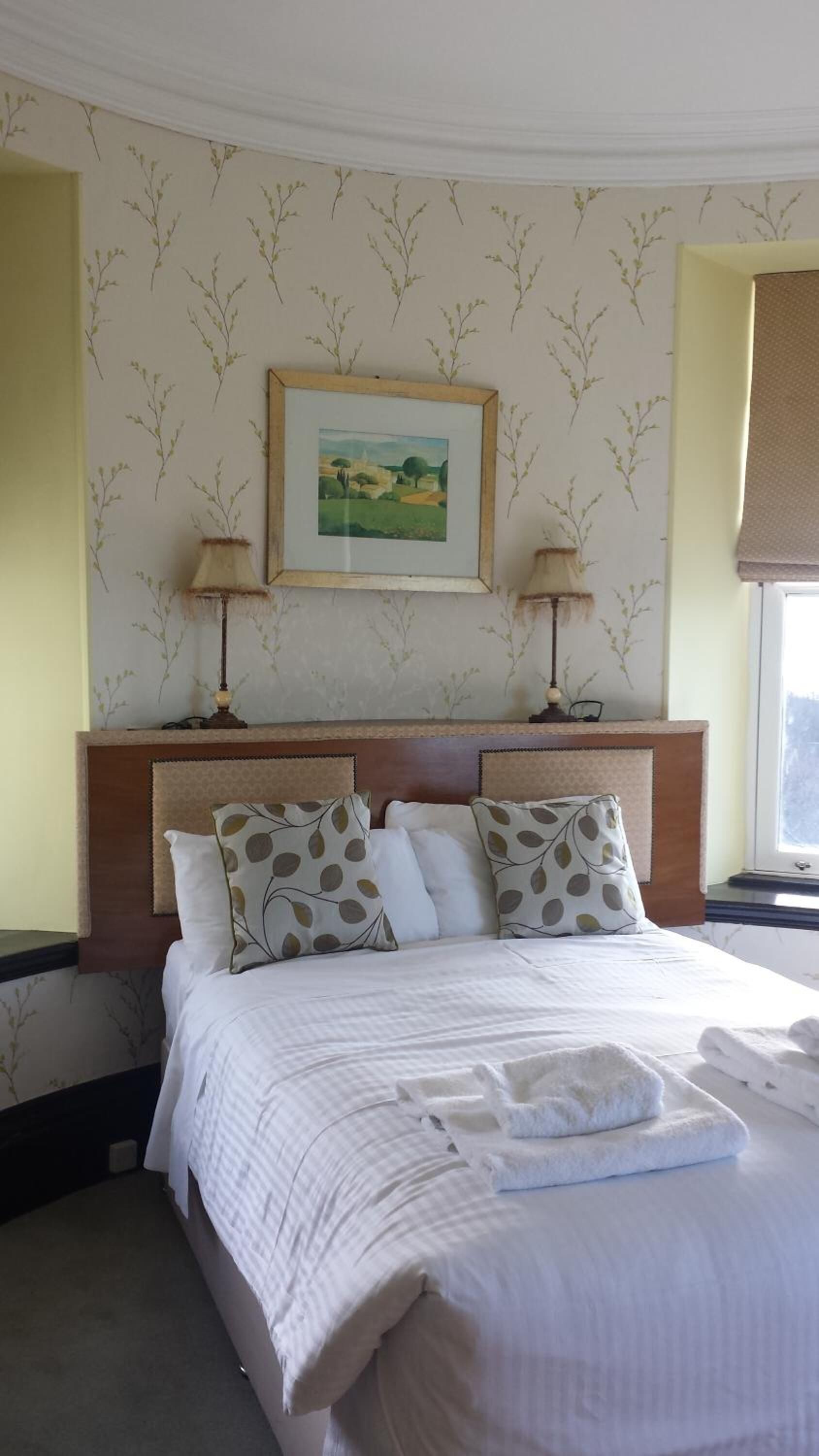 A hotel room with a large bed, two bedside lamps, a framed landscape painting, and a window with a view.