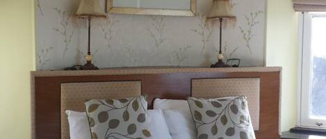 A hotel room with a large bed, two bedside lamps, a framed landscape painting, and a window with a view.