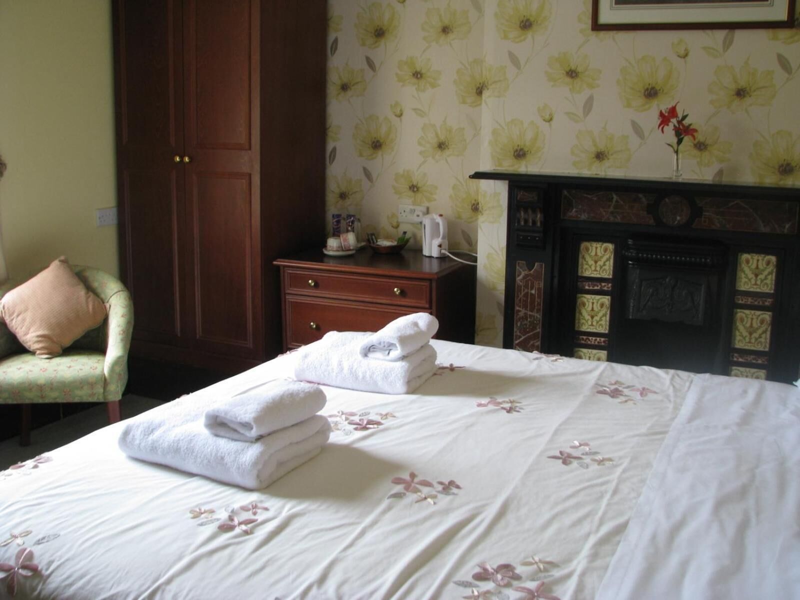 A hotel room with a bed, a wooden dresser, a green armchair, and a fireplace.
