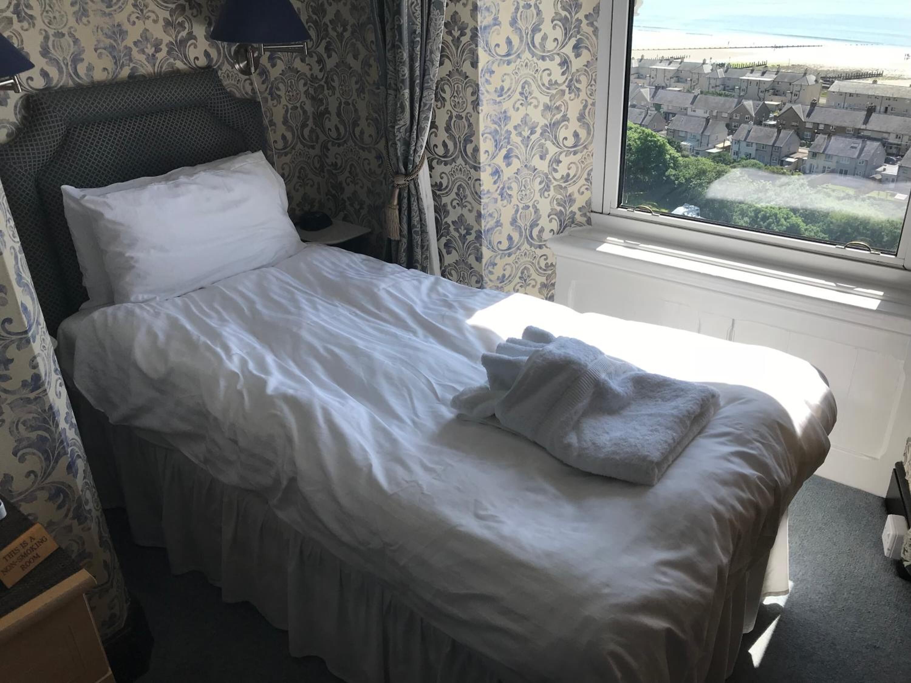 Ein ordentlich gemachtes Bett mit weißen Bettwäsche und einer grauen Decke, eine Nachttischlampe und ein Fenster mit Blick auf eine Wohngegend.