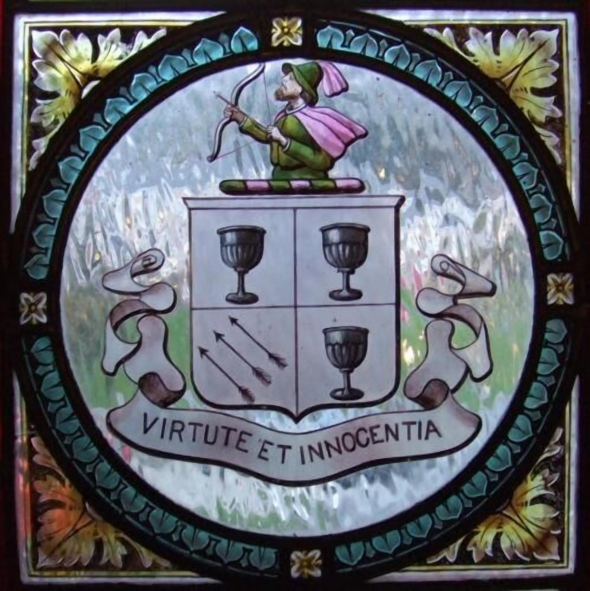 Une fenêtre en vitrail ornée d’un blason représentant une figure sur un écu, deux lions et la devise « VIRTUTE ET INNOCENTIA ».