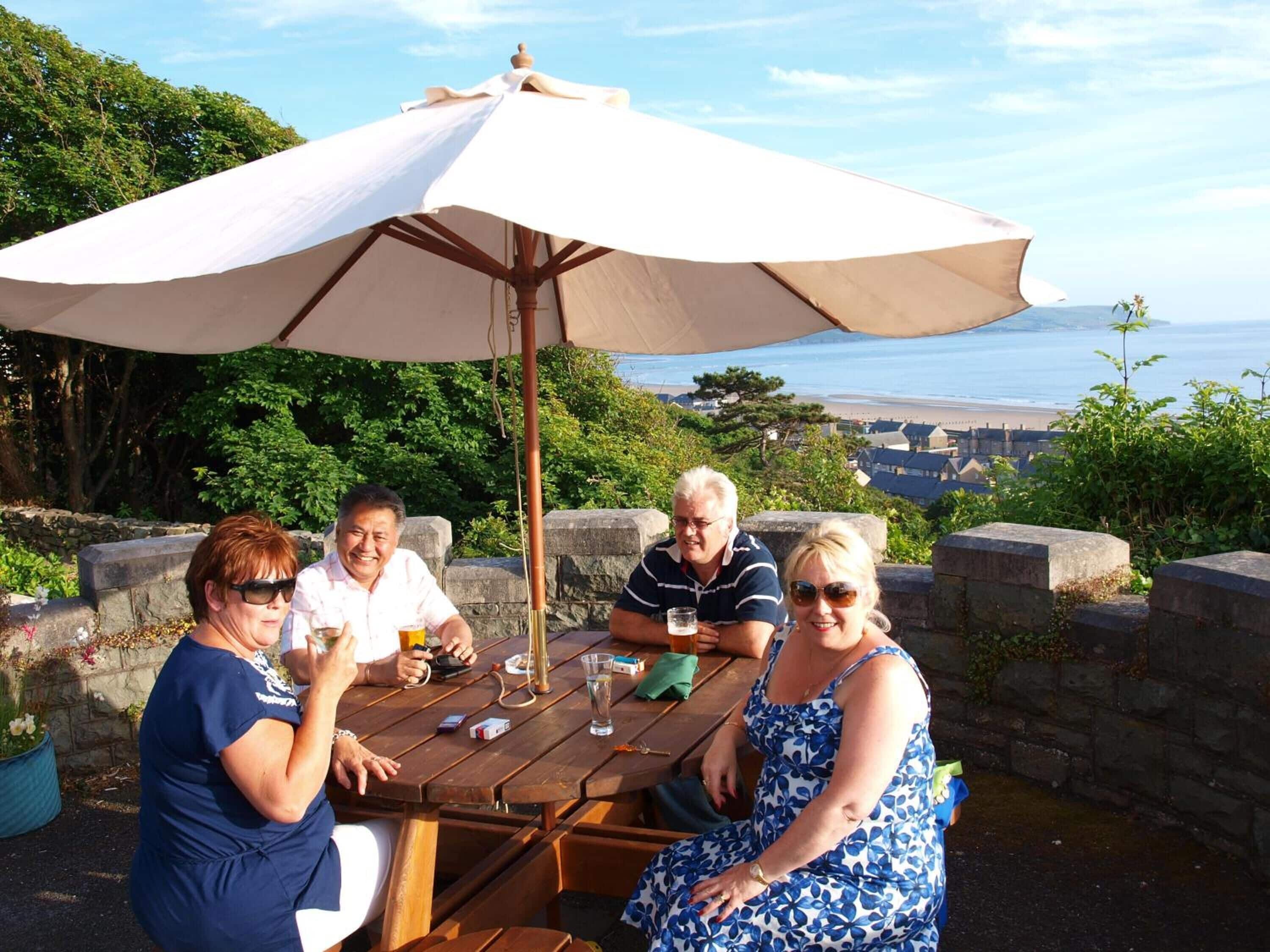 Een groep mensen zit aan een houten tafel onder een grote parasol, met uitzicht op een prachtig kustlandschap.