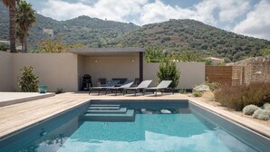 Una piscina con lettini, una zona relax coperta e uno sfondo montuoso.