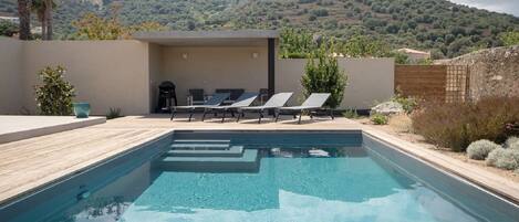A swimming pool with lounge chairs, a covered seating area, and a mountainous backdrop.