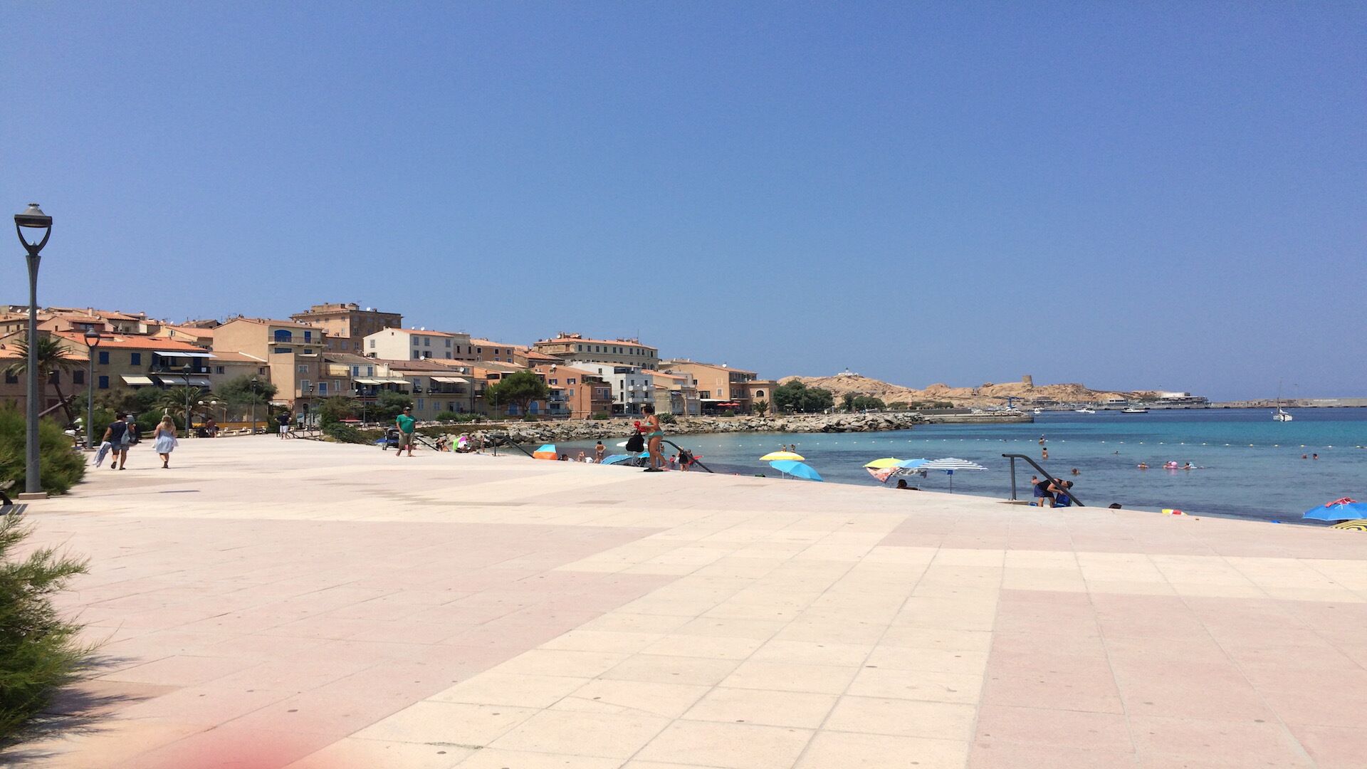 A coastal promenade with a clear view of the sea, a tiled walkway, and buildings in the background.