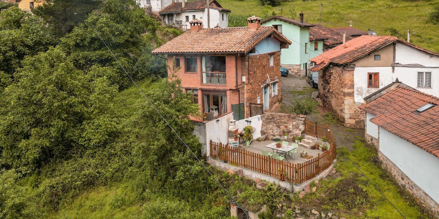 Un pueblo en una ladera con casas tradicionales, un patio central con una mesa y sillas, y un muro de piedra.