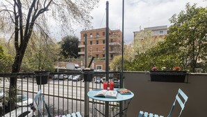 A balcony with a table and chairs, a view of a street, and a building in the background.
