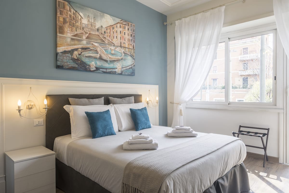A neatly made bed with white and blue bedding, a painting of a canal scene above it, and a window with curtains to the right.