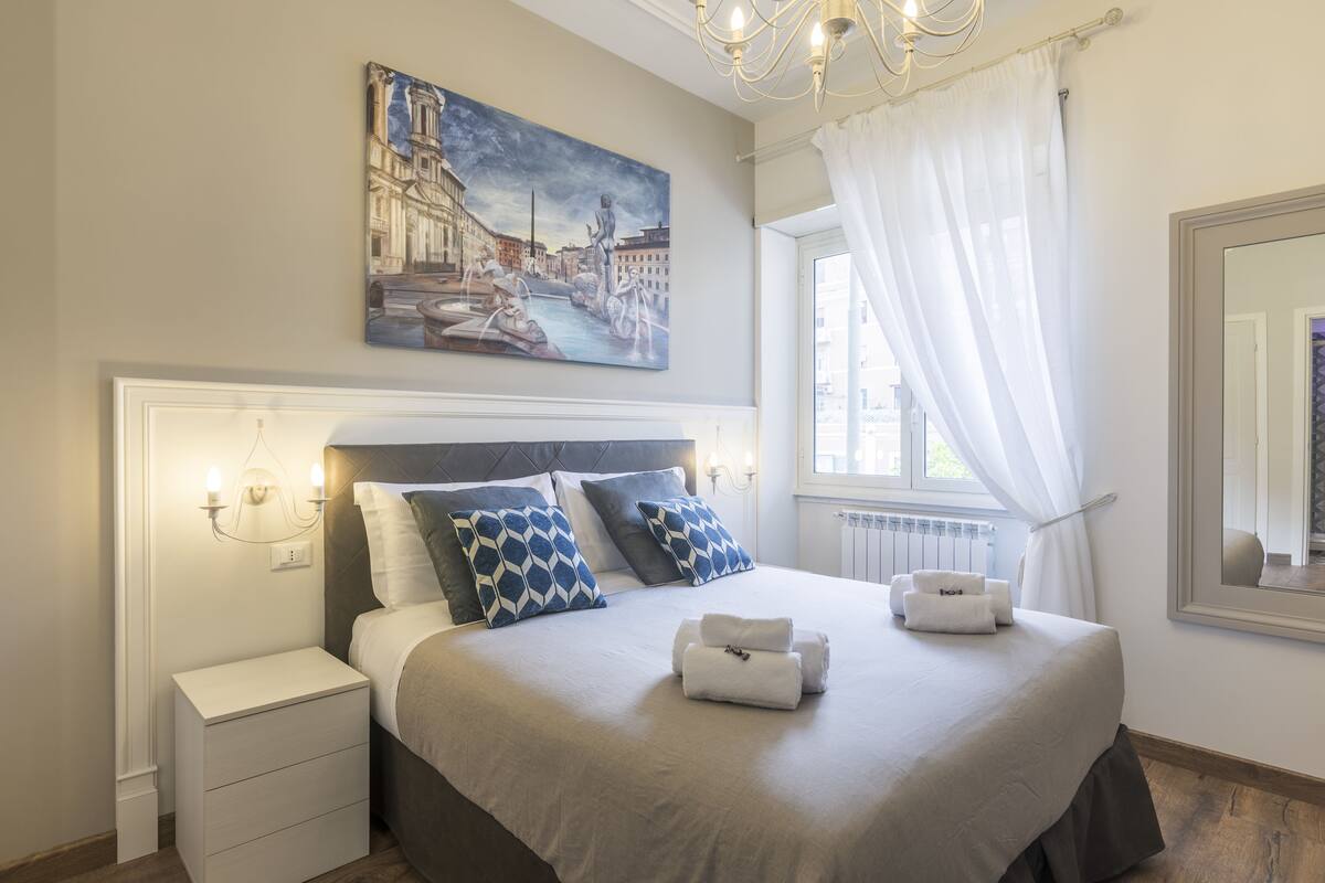 A neatly arranged bedroom with a large bed, a nightstand, a painting of a cityscape, and a window with curtains.
