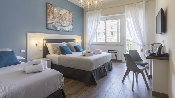A hotel room with two beds, a desk, and a chair. There is a painting on the wall, a chandelier, and a window with curtains.