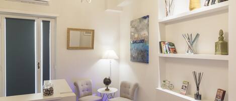 Habitación de hotel con un mostrador de recepción blanco, dos sillones, una mesita redonda pequeña, un globo terráqueo, una lámpara, un cuadro y estantes con libros y objetos decorativos.