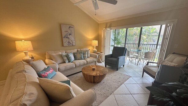 A living room with a beige sofa, a coffee table, and a chair. There is a ceiling fan, a painting on the wall, and a window with a view of greenery outside. - Enjoy the tranquility of this spacious two bedroom unit. Siesta Dunes 6-203 (Siesta Key)