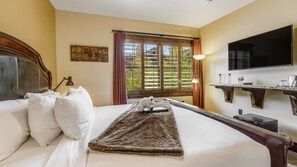 Quarto de hotel com cama, televisão, janela com cortinas, quadro na parede e uma pequena mesa com objetos.