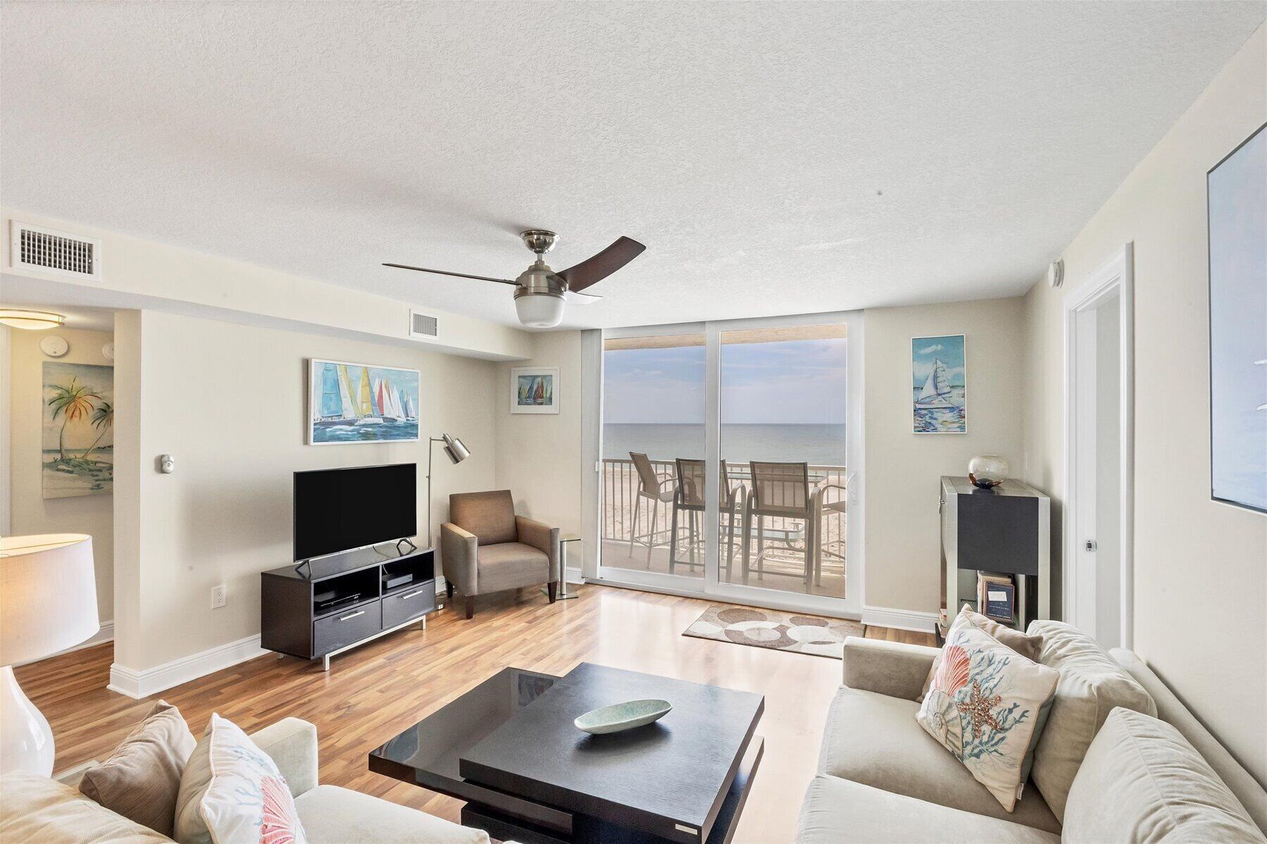Een ruime woonkamer met een flatscreen-tv, comfortabele zithoek, een plafondventilator en een grote schuifpui die leidt naar een balkon met uitzicht op het strand.