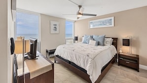 Quarto com cama, mesas de cabeceira, ventilador de teto e uma grande janela com vista para o mar.