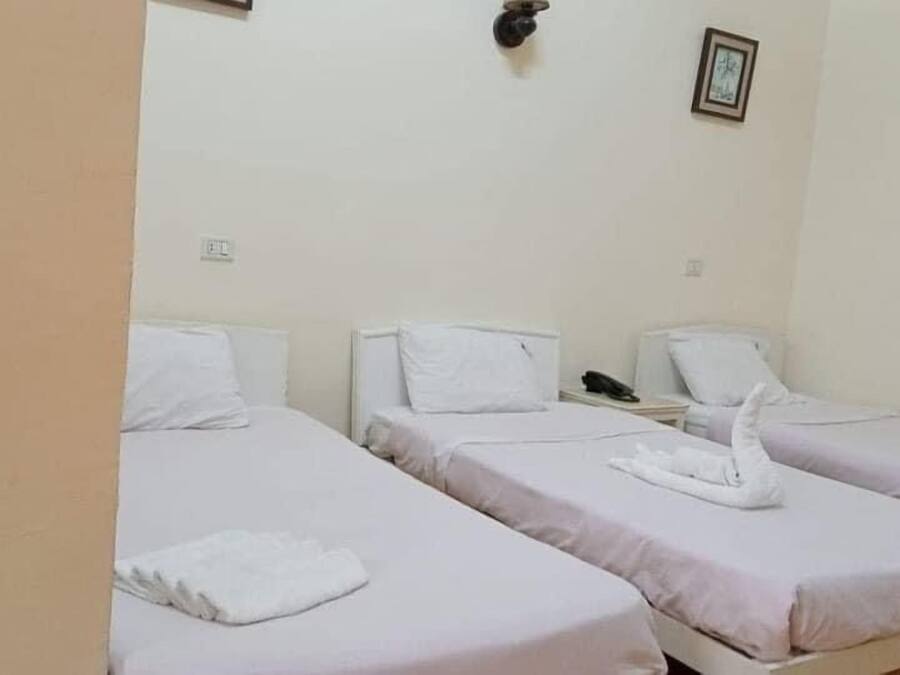 Une chambre avec trois lits simples, chacun doté de draps et d’oreillers blancs. On y trouve une petite table avec un téléphone et un tableau encadré accroché au mur.