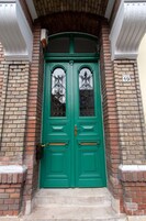 Una puerta doble verde con un pequeño ventanal encima, ubicada en un edificio de ladrillo con un dintel blanco y una pequeña placa a la derecha.