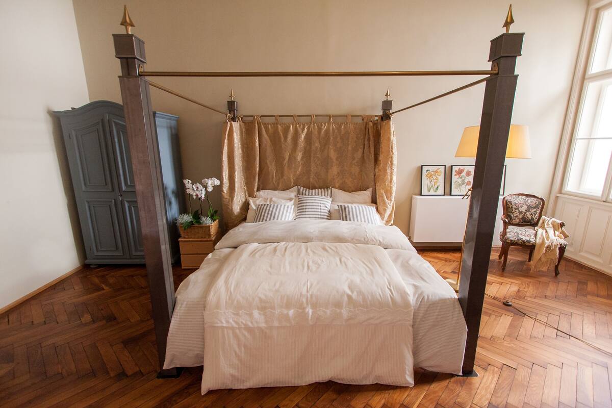 Una cama con dosel sobre pilares, suelo de madera, una mesita de noche con lámpara, una silla y un armario.