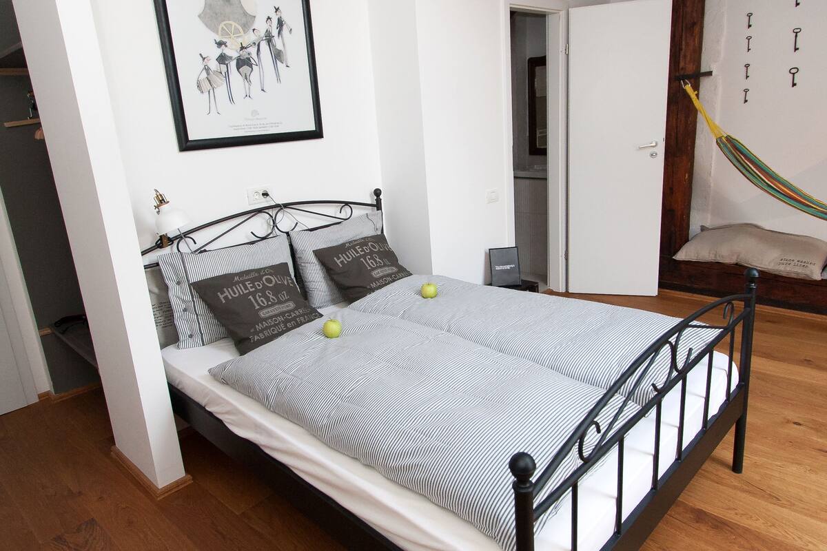 Un dormitorio con cama, suelo de madera, un cuadro enmarcado en la pared y una lámpara colgante.