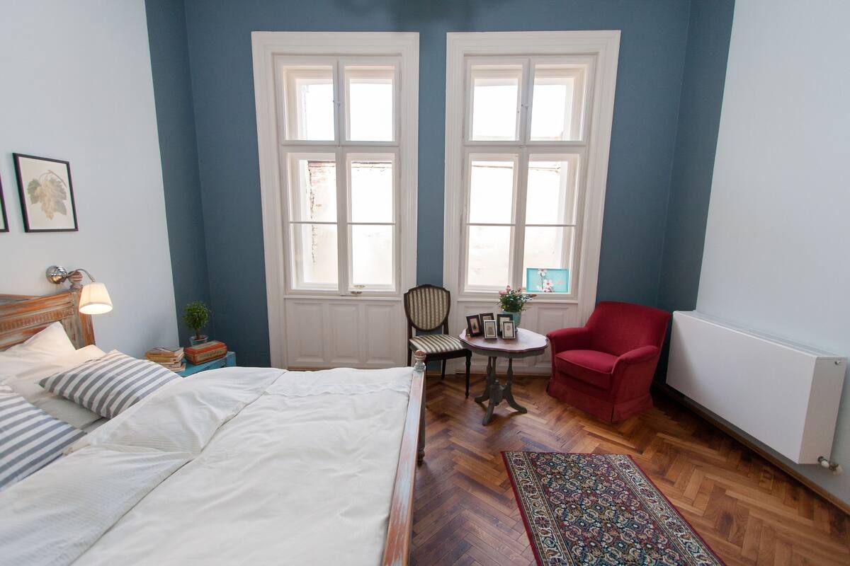 Un dormitorio con una cama, dos ventanas, una silla, una mesita y un sillón rojo.
