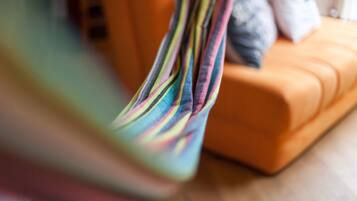 A close-up of a colorful striped fabric, possibly a hammock or a piece of clothing, with a blurred background featuring an orange couch and a white pillow.